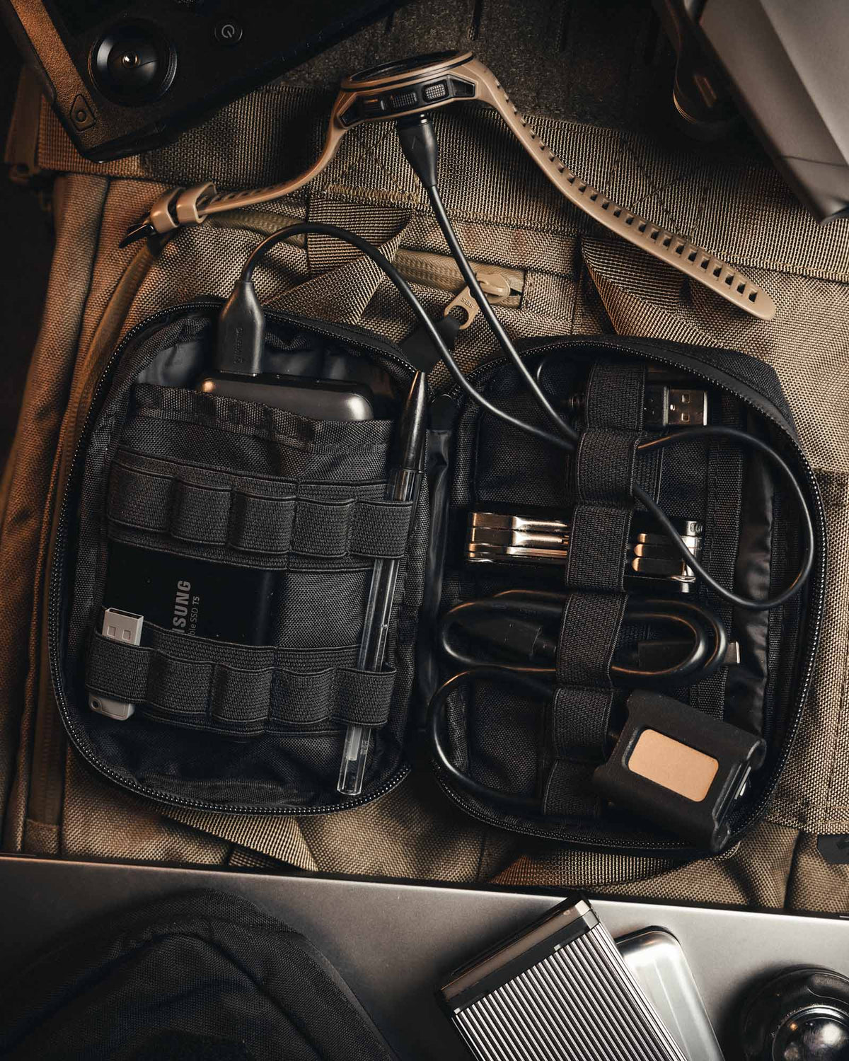 Die geöffnete STOIRM Large Pouch V.1 in Olive Green von STOIRM bietet Platz für Kabel, eine tragbare SSD von Samsung, einen USB-Adapter, Stifte und anderes technisches Zubehör auf einer hellbraunen Stoffoberfläche mit Molle-Panel.