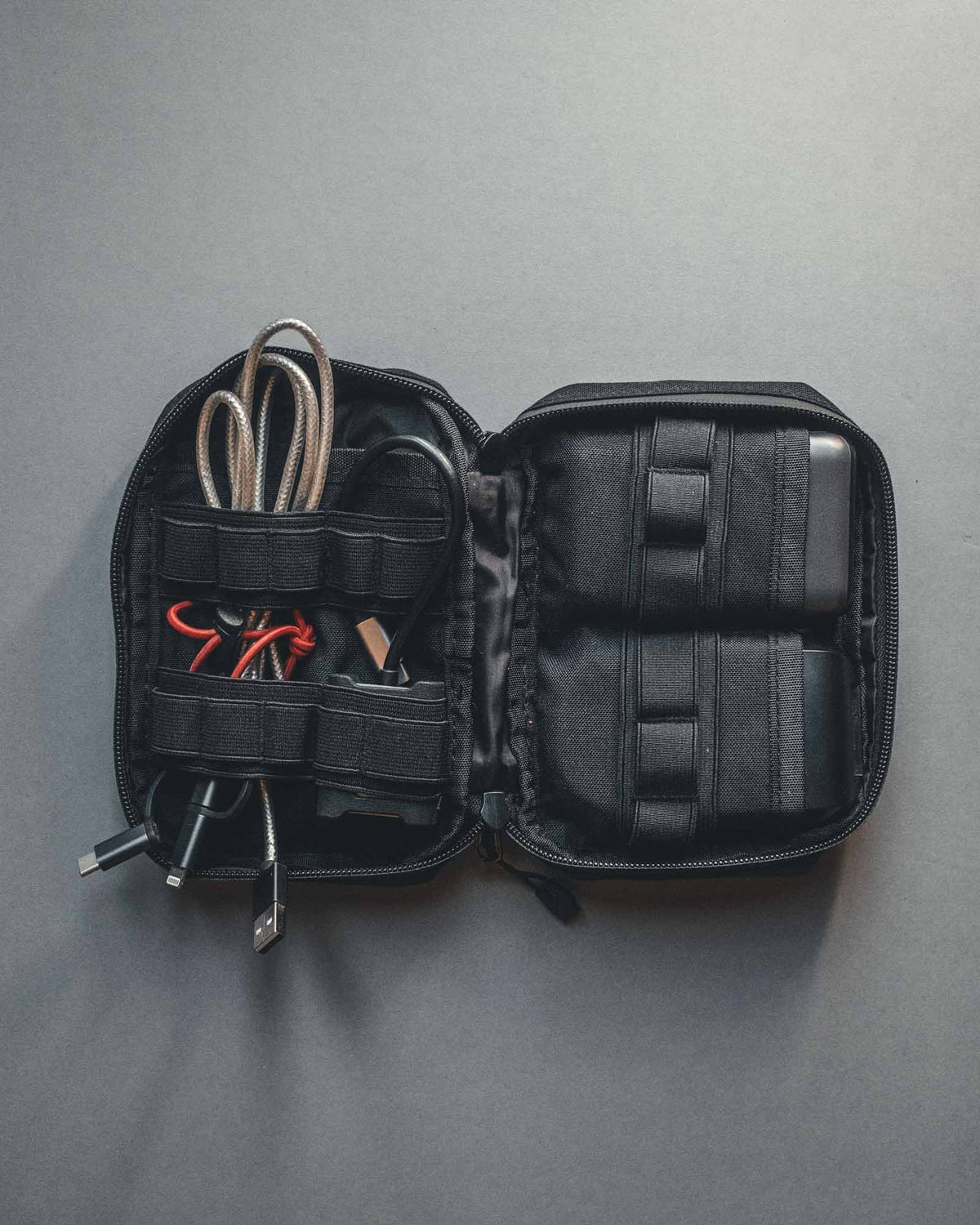 In der offenen STOIRM Large Pouch V.1 von STOIRM in Olivgrün sind Ladekabel, ein USB-Stick und zwei schwarze elektronische Zubehörteile in elastischen Fächern vor einem grauen Hintergrund ordentlich untergebracht.