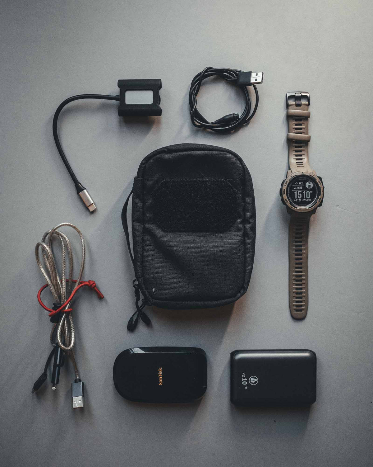 Eine flache Anordnung von Gadgets und Zubehör auf einer grauen Oberfläche, mit einer Smartwatch, einer olivgrünen STOIRM Large Pouch V.1 von STOIRM mit Molle-Panel, Ladekabeln, einem USB-Adapter, einem SanDisk-Gerät und einer schwarzen Powerbank.