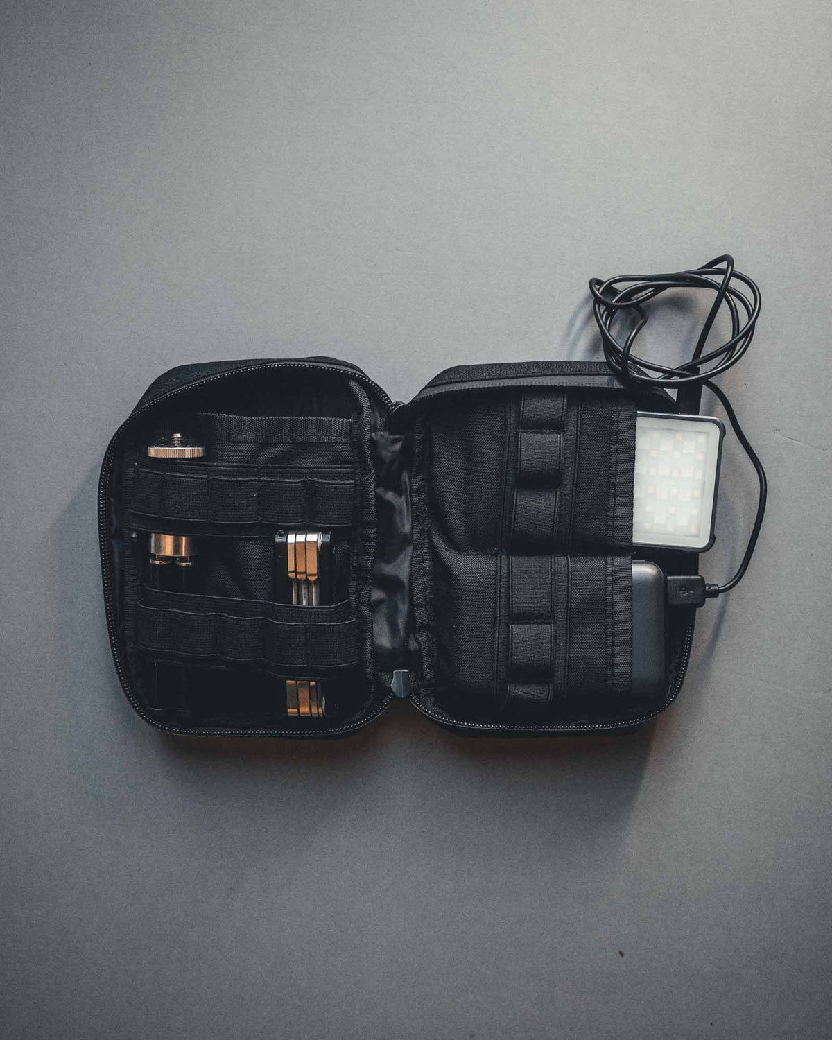 Ein schwarzer STOIRM Large Pouch V.1 Tech-Organizer hält Kabel, Gadgets und eine rechteckige LED-Lampe in elastischen Bändern und Taschen, offen gezeigt auf einer grauen Oberfläche neben einem olivgrünen Molle-Panel.