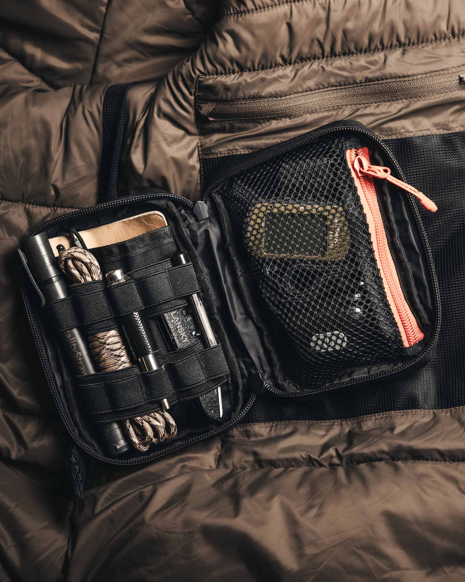 Ein kompaktes Survival-Kit in einer STOIRM Large Pouch V.2 von STOIRM liegt auf einer braunen, gesteppten Oberfläche. Auf der linken Seite befinden sich eine Taschenlampe, ein Paracord, ein Stift und Werkzeuge, auf der rechten Seite ein kleines gelb-schwarzes Gerät in der Netztasche mit Reißverschluss.