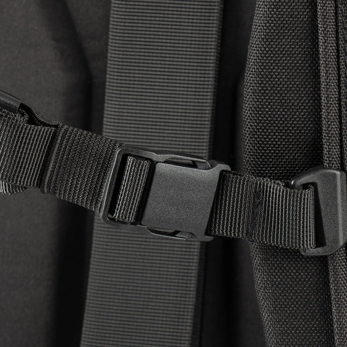 Nahaufnahme einer schwarzen Kunststoffschnalle, die an zwei verstellbaren Nylonriemen befestigt ist. Sie ist Teil der STOIRM Messenger Bag von STOIRM, die für Laptops bis zu 17 Zoll geeignet ist und vor einem Hintergrund aus strukturiertem Stoff gezeigt wird.