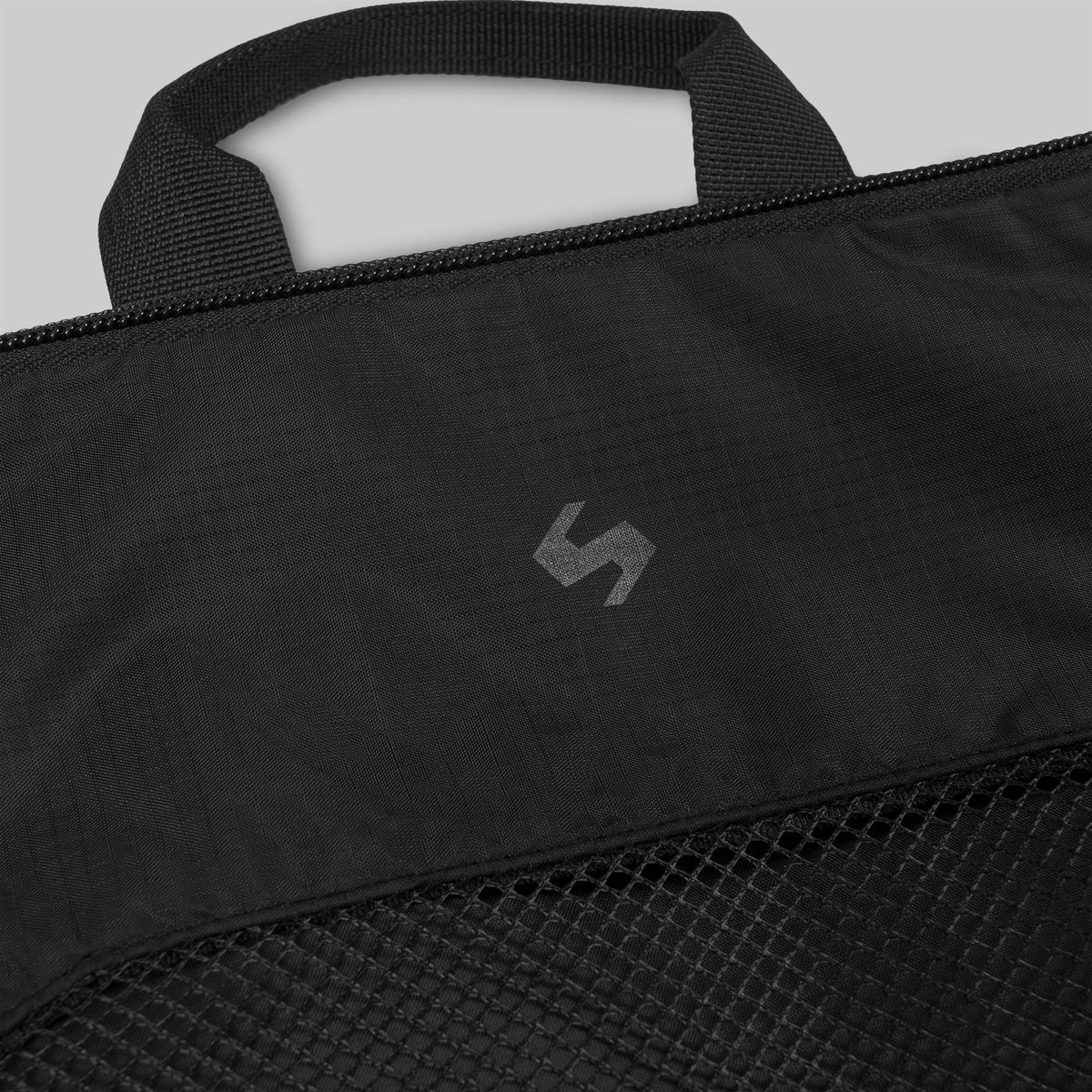 Nahaufnahme des STOIRM Travel Cube with Laundry Bag von STOIRM mit schwarzem Stoff, Netztasche, kleinem Griff, Reißverschluss und einem dezenten grauen, geometrischen Logo auf der Oberfläche.