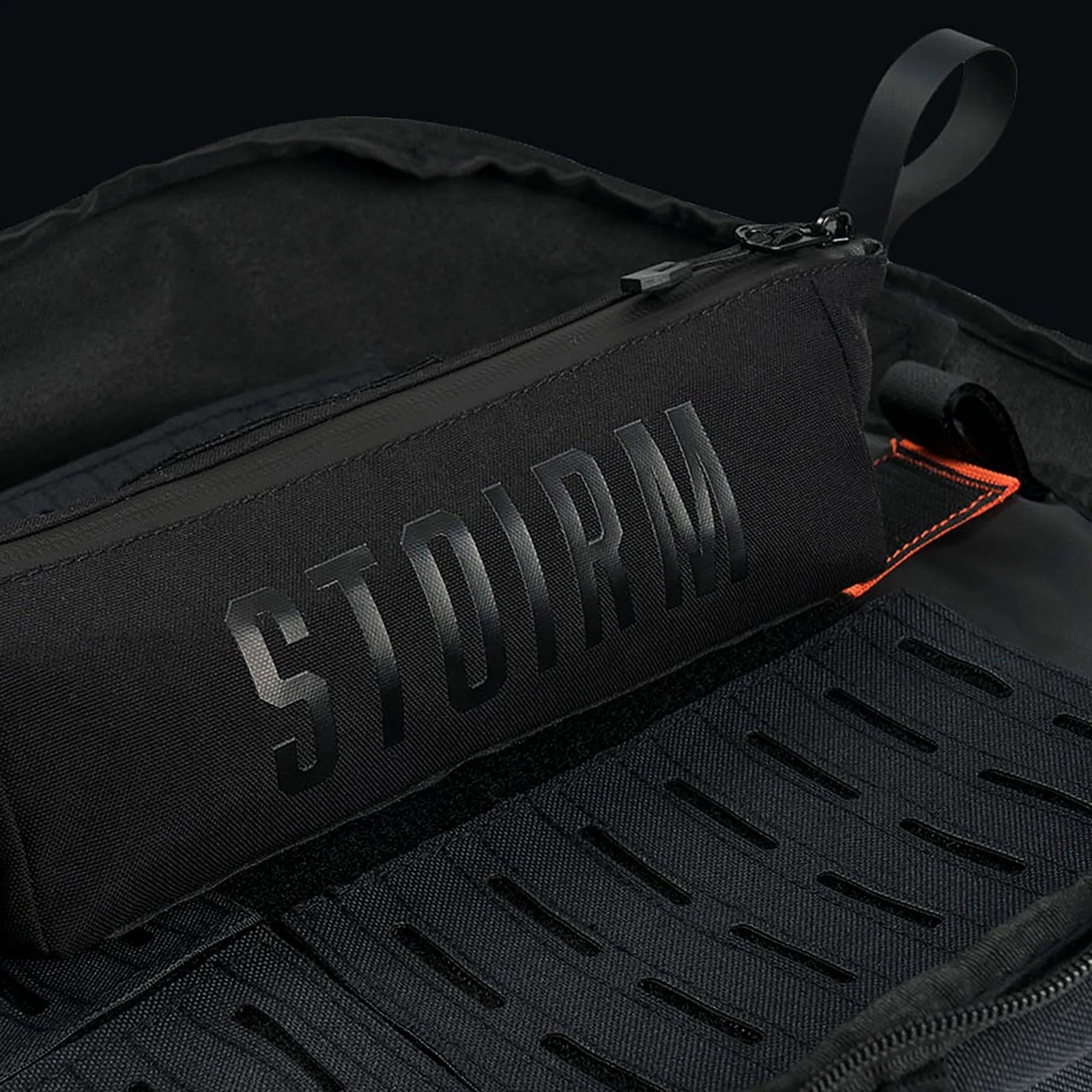Eine schwarze STOIRM Utility Pouch mit auffälligem Branding befindet sich in einem schwarzen, wasserdichten Beutel mit offenem Reißverschluss, der strukturierte Gittereinsätze und einen kleinen orangefarbenen Akzent offenbart.
