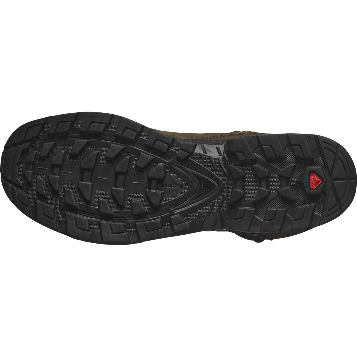 Das Bild zeigt die schwarze, strukturierte Außensohle des SALOMON TRACKER QUEST TRACKER HIGH GTX in Earth Brown/Green mit tiefen, multidirektionalen Stollen für die Traktion und einem kleinen roten Dreieckslogo im Zehenbereich.