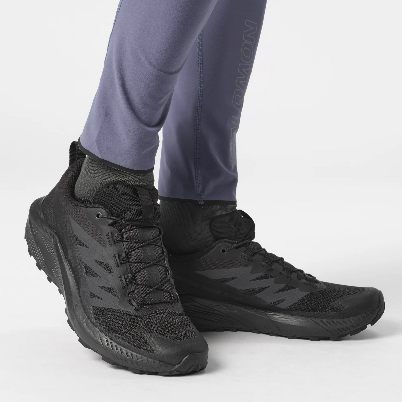 Eine Person in lilafarbenen Leggings mit sichtbarem Unterschenkellogo trägt schwarze SALOMON FORCES SENSE RIDE 5 SR Schuhe mit All Terrain Contagrip Sohlen, stehend auf weißem Hintergrund.
