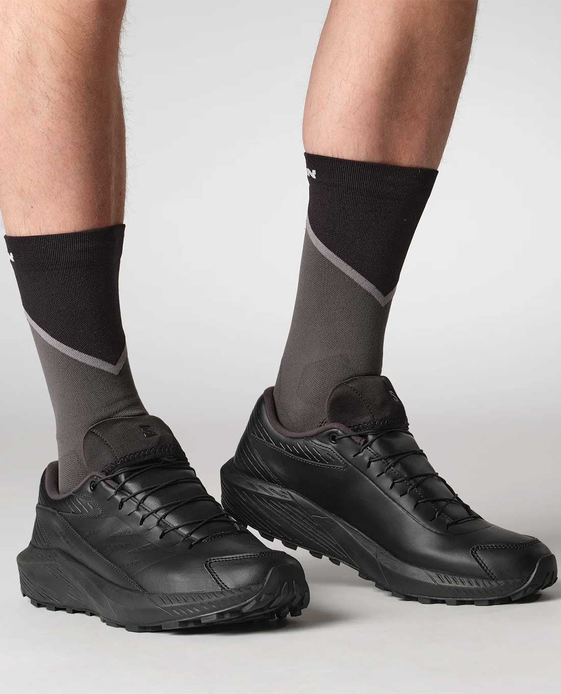 Mit SALOMON FORCES Salomon SENSE RIDE 5 SR LEATHER - Black und zweifarbigen schwarz-grauen Socken steht eine Person auf einer hellgrauen Fläche. Die robusten Sohlen suggerieren Outdoor-Abenteuer, wobei nur die Unterschenkel sichtbar sind.