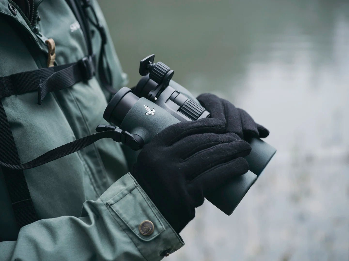 Eine Person in grüner Jacke und schwarzen Handschuhen hält eine SWAROVSKI OPTIK NL Pure mit Swarovski FRP Stirnstütze in der Nähe eines ruhigen Gewässers, bereit, die Szenerie zu beobachten.