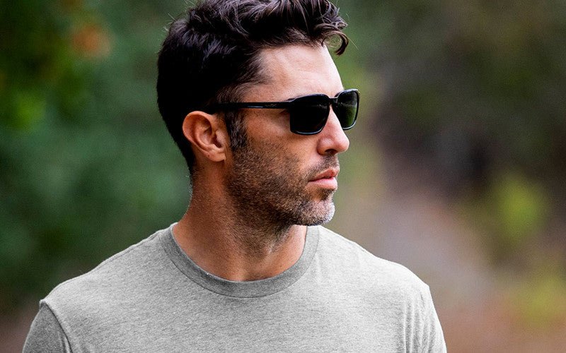 Ein Mann in einem hellgrauen T-Shirt und einer WILEY X Sonnenbrille ALFA mit grauen Gläsern steht im Freien und schaut zur Seite vor einem unscharfen grünen und braunen Naturhintergrund.