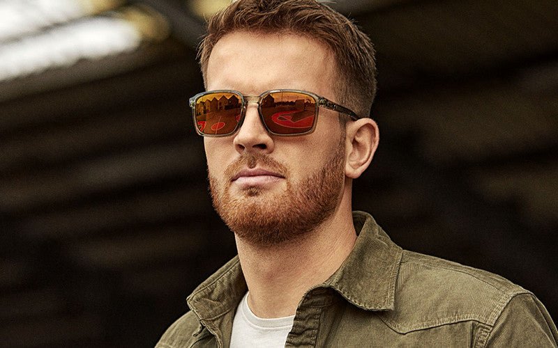 Ein Mann mit kurzen roten Haaren und einem Bart trägt eine WILEY X Sonnenbrille ALFA mit abnehmbarem Seitenschutz und eine grüne Jacke über einem weißen Hemd. Er steht im Freien mit verschwommenen Strukturen im Hintergrund.