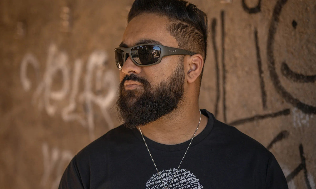 Ein bärtiger Mann, der eine WILEY X CLIMB Sonnenbrille mit 100% UVA/UVB-Schutz und ein schwarzes T-Shirt trägt, steht vor einer mit Graffiti bedeckten Wand. Die Schatten in seinem Gesicht heben den lebendigen, strukturierten Hintergrund hervor.