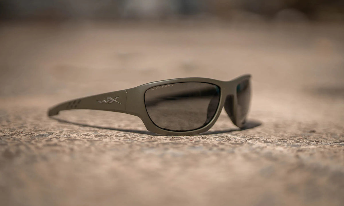Eine WILEY X Wiley X Sonnenbrille CLIMB in OD Green mit dunklen Gläsern und 100% UVA/UVB-Schutz ruht auf einer strukturierten, sandigen Oberfläche vor einem verschwommenen Hintergrund.