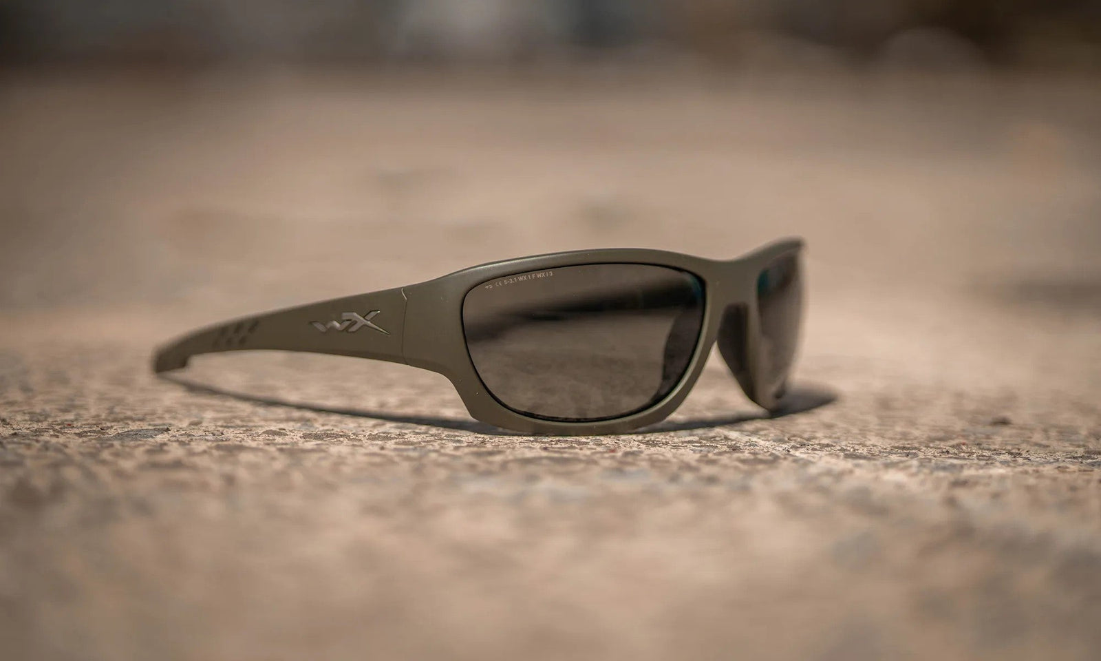 Eine WILEY X Wiley X Sonnenbrille CLIMB in OD Green mit dunklen Gläsern und 100% UVA/UVB-Schutz ruht auf einer strukturierten, sandigen Oberfläche vor einem verschwommenen Hintergrund.