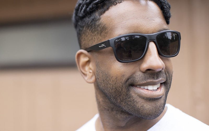 Ein Mann mit kurzen lockigen Haaren und gestutztem Bart lächelt, während er eine WILEY X OVATION Sonnenbrille und ein weißes Hemd trägt und im Freien vor einem unscharfen braunen Hintergrund steht.