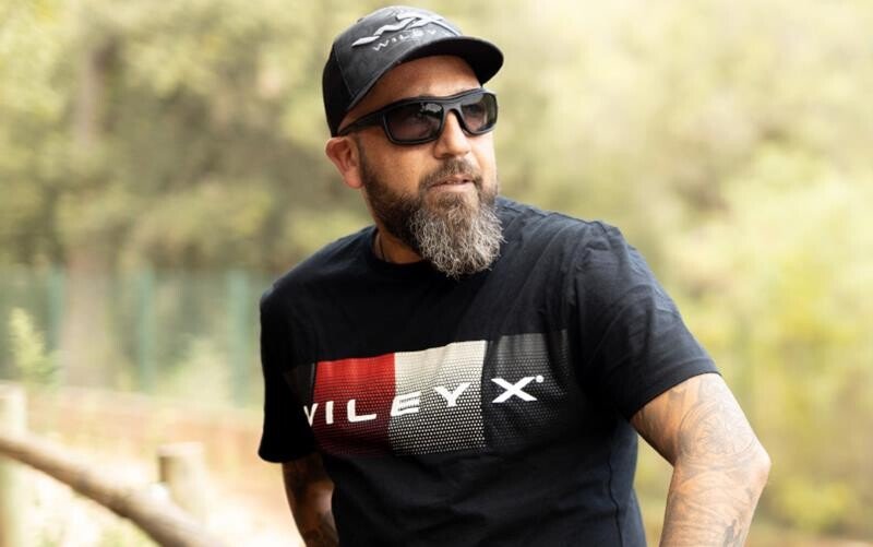 Ein bärtiger Mann, der eine Wiley X Sonnenbrille OZONE, eine schwarze Kappe und ein schwarzes T-Shirt mit dem WILEY X-Logo trägt, steht im Freien mit verschwommenen Bäumen und einem Zaun im Hintergrund.