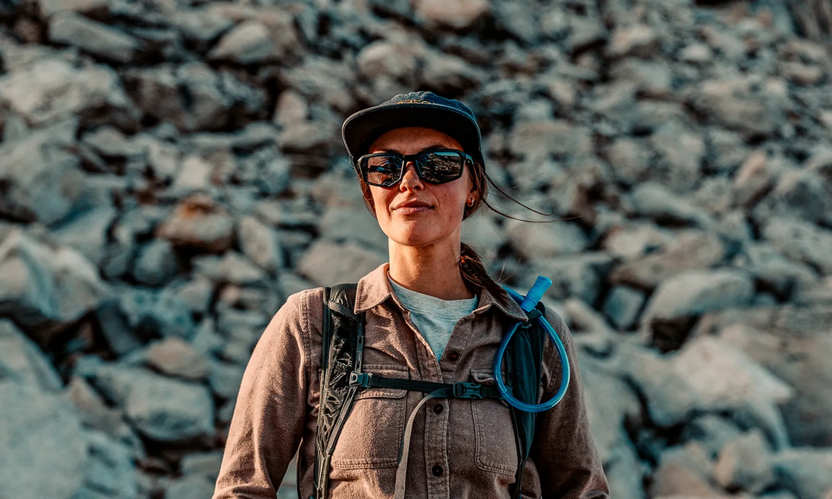 Eine Frau in Outdoor-Wanderkleidung steht selbstbewusst vor einem felsigen Berghang und trägt einen WX Recon Trinkrucksack, eine Mütze und die WILEY X Sonnenbrille RECON mit CAPTIVATE Gläsern.