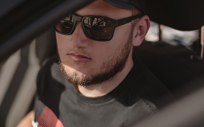 Ein Mann mit kurzem Bart und Schnurrbart, der eine dunkle Sonnenbrille - speziell die WILEY X Sonnenbrille TREK -, ein schwarzes T-Shirt und eine Kappe trägt, sitzt auf dem Fahrersitz eines Autos und schaut leicht zur Seite.