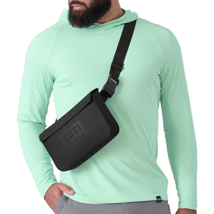 Eine Person in einem hellgrünen, langärmeligen Kapuzenpulli und einer grauen Hose steht mit einer schwarzen YETI Crossbody Bag mit dem YETI® Sideclick® Gurt - Black, einem verstellbaren und strapazierfähigen Nylongurt von YETI, auf der Brust.