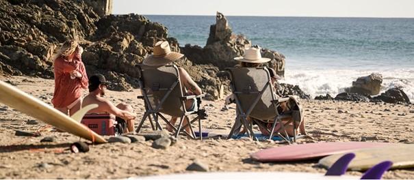 Vier Personen entspannen auf YETI® Camping Stuhl Trailhead - Navy Stühlen und einer Kühlbox an einem felsigen Strand mit Blick auf den Ozean. Zwei Surfbretter liegen in der Nähe, während sie die Aussicht auf das Meer genießen.