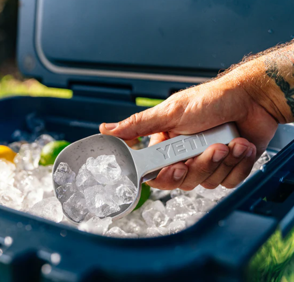 Eine Hand hält einen mit Eis gefüllten YETI® Eislöffel über eine offene Kühlbox, die mit Eis, Zitronen und Limetten gefüllt ist; diese spülmaschinenfeste Schaufel von YETI ist perfekt für jede Zusammenkunft.