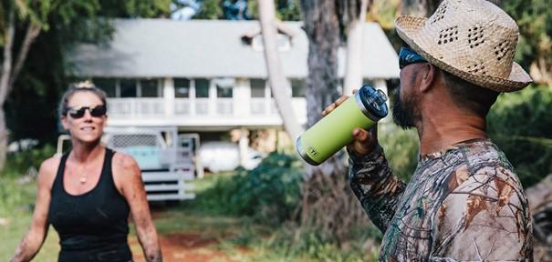 Eine Frau in einem schwarzen Tank-Top lächelt neben einem Mann mit Strohhut und Tarnhemd und trinkt aus einer weißen YETI® Flasche Rambler 18 Oz mit doppelwandiger Vakuumisolierung. Im Freien, mit einem Haus und Grünflächen im Hintergrund.