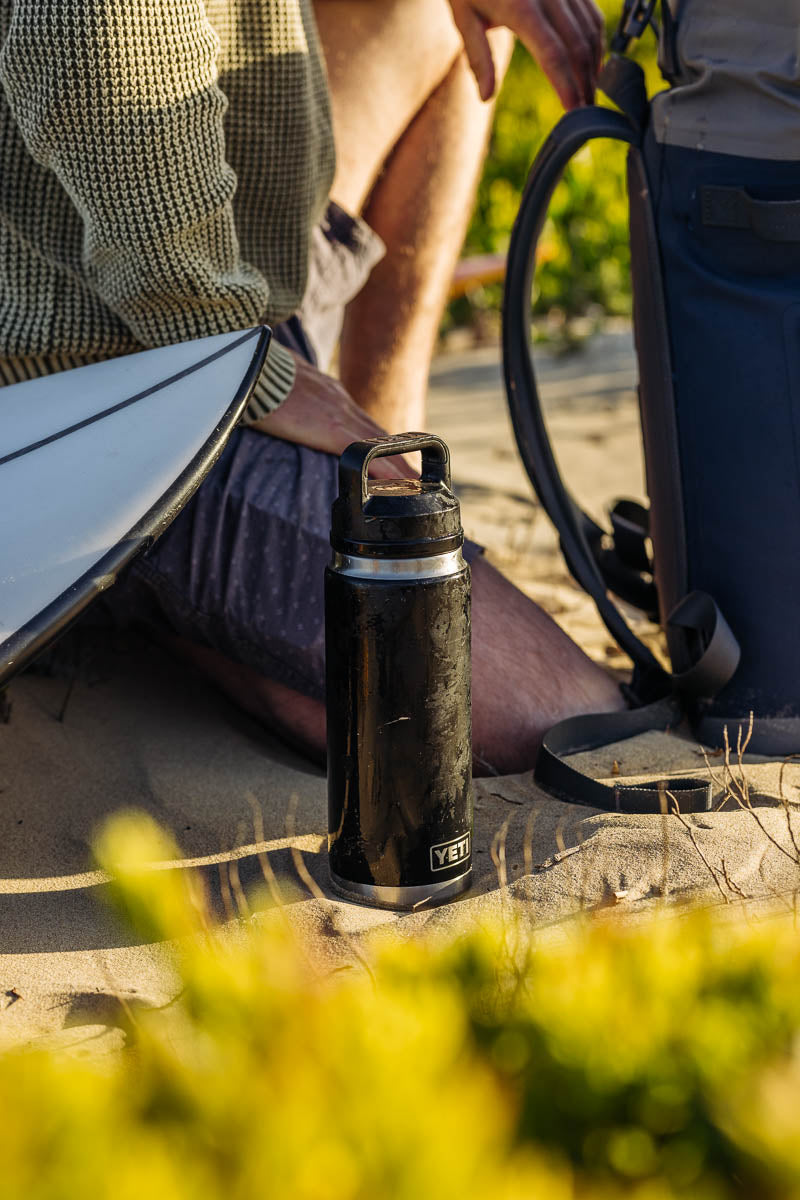 Eine YETI® Flasche Rambler 26 Oz - Black sitzt auf sandigem Boden in der Nähe einer Person in Pullover und Shorts, mit einem Surfbrett, einem blauen Rucksack und grünem Laub in der Nähe.