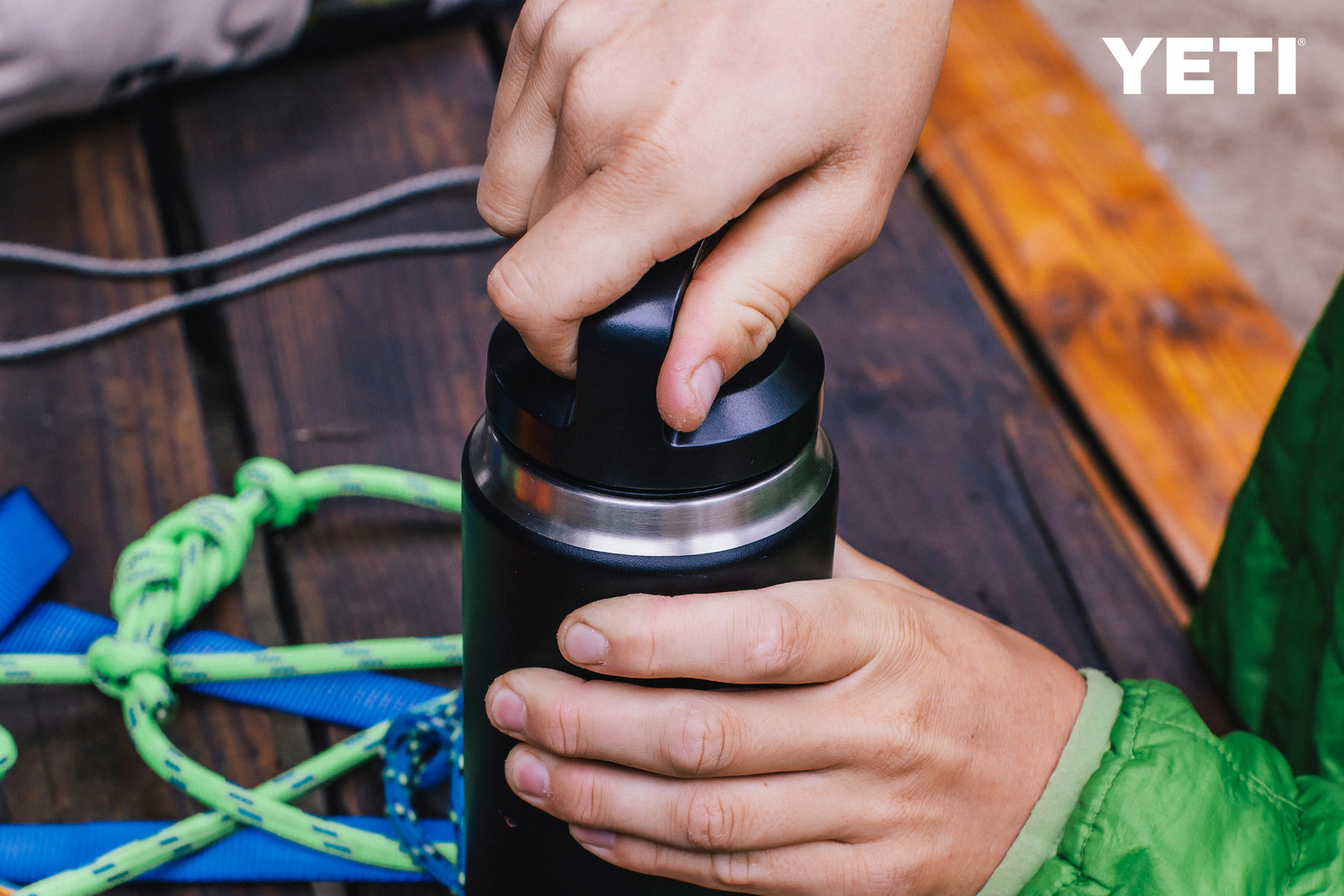 Eine Person in grüner Jacke öffnet eine schwarze YETI® Flasche Rambler 26 Oz auf einer hölzernen Oberfläche mit Kletterausrüstung in der Nähe. Die doppelwandige Vakuumisolierung hält Getränke kalt, und das YETI Logo ist oben rechts sichtbar.