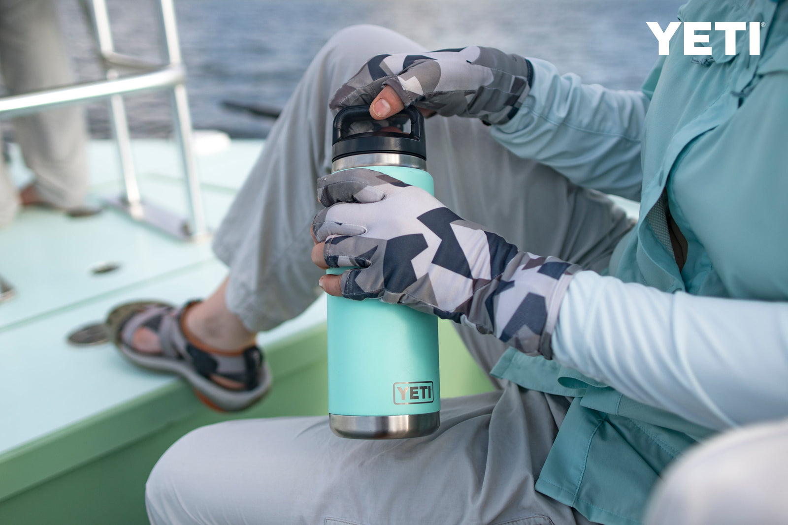 Ein Angler mit Anglerhandschuhen und Sonnenschutzkleidung öffnet eine marineblaue YETI® Flasche Rambler 26 Oz mit auslaufsicherem Strohdeckel, während er auf einem Bootsdeck sitzt, im Hintergrund ist Wasser zu sehen.
