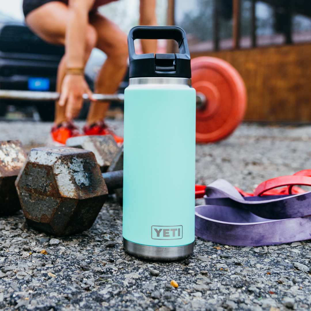 Eine navyfarbene YETI® Flasche Rambler 26 Oz Straw steht auf Kies neben Hanteln, Widerstandsbändern und einer geladenen Langhantel, während sich jemand darauf vorbereitet, im Freien Gewichte zu stemmen.