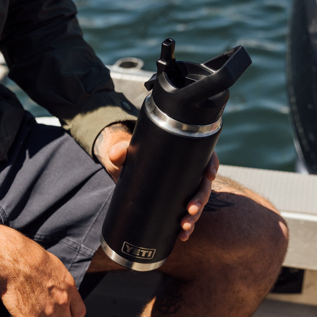 Eine Person sitzt auf einem Boot und hält eine YETI® Flasche Rambler 26 Oz Straw in Seeschaum. Das ruhige Wasser ist im Hintergrund, und sie trägt graue Shorts und eine grüne Jacke.