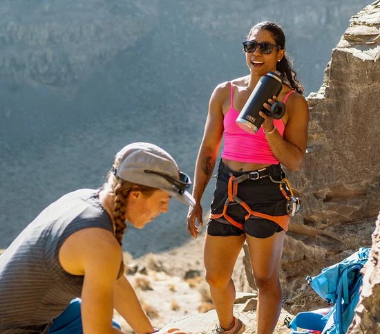 Zwei Frauen in Kletterausrüstung stehen auf einem felsigen Vorsprung. Die eine steht lächelnd mit einer spülmaschinenfesten YETI® Rambler 36 Oz Flasche in Rescue Red in der Hand, während die andere sich hinkniet, um ihre Ausrüstung anzupassen. Das Sonnenlicht beleuchtet den Canyon hinter ihnen.