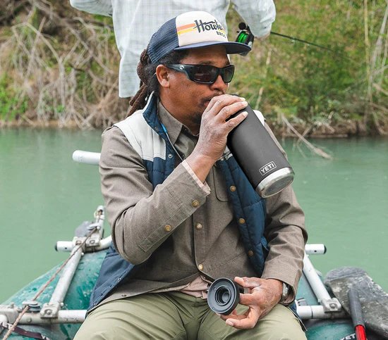 Ein Mann mit Sonnenbrille, Mütze und Outdoor-Kleidung sitzt in einem Boot auf einem Fluss und trinkt aus einer spülmaschinenfesten YETI® Flasche Rambler 36 Oz in Rescue Red. Eine andere Person steht hinter ihm und hält eine Angelrute.
