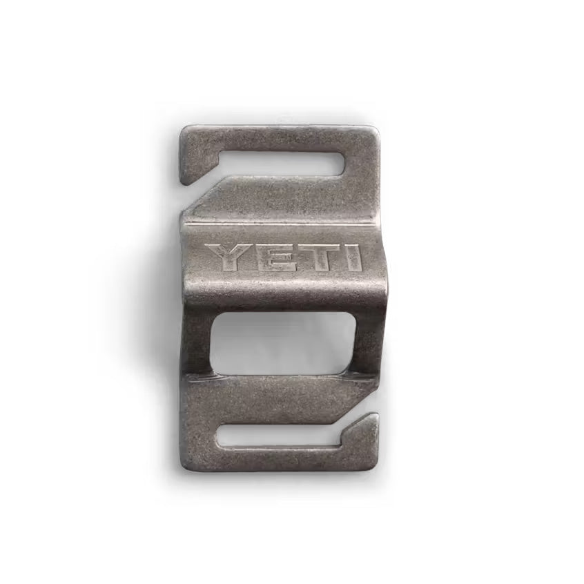 Der YETI® Flaschenöffner - Mollele Bottle Opener ist ein silberner Edelstahl-Clip mit rechteckigen Ausschnitten, hakenförmigen Kanten und einem geprägten YETI-Logo. Kompatibel mit dem MOLLE-System. Marke: YETI. Abgebildet auf weißem Hintergrund.
