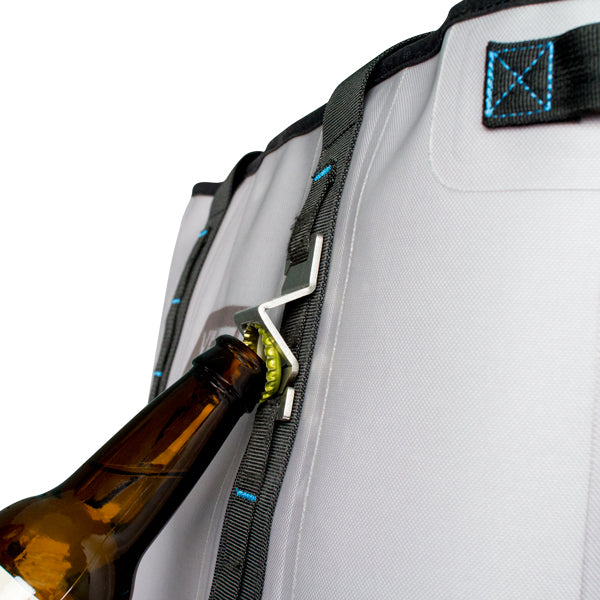 Nahaufnahme einer Bierflasche, die mit dem YETI® Flaschenöffner - Mollele Bottle Opener aus rostfreiem Stahl geöffnet wird. Der Flaschenöffner ist am Gurt des MOLLE-Systems eines grauen Rucksacks mit schwarzen und blauen Nähten befestigt.