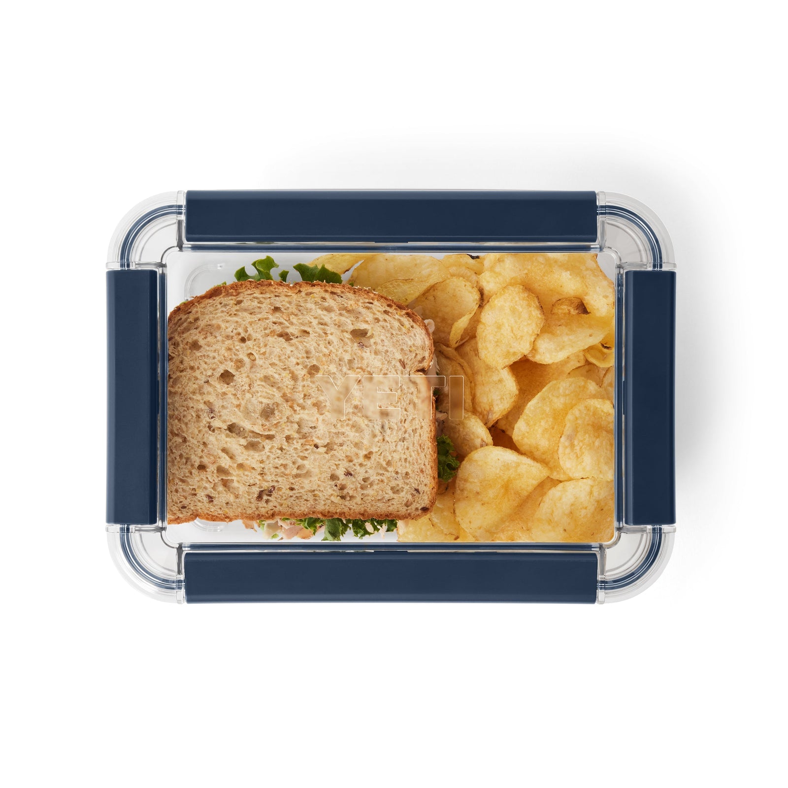 Ein klarer, rechteckiger YETI® Food Storage Large von YETI fasst ein Vollkornsandwich und Kartoffelchips, von oben betrachtet. Der BPA-freie Behälter hat dunkelblaue Griffe und eignet sich perfekt für die Aufbewahrung von Lebensmitteln für unterwegs.
