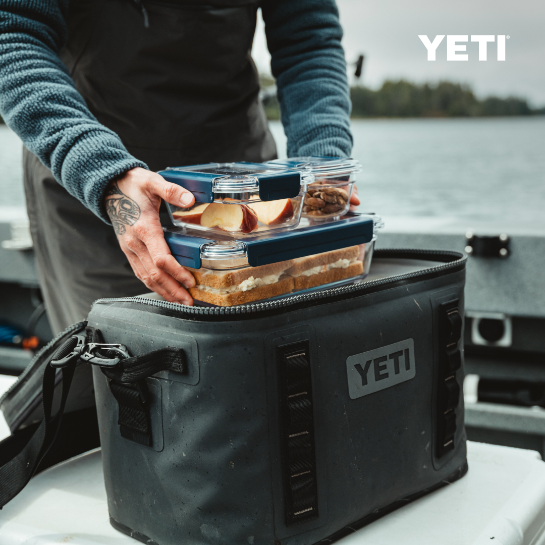 Jemand stellt zwei BPA-freie YETI® Food Storage Small-Behälter in eine schwarze YETI-Kühltasche auf einem Boot, mit Wasser und Bäumen dahinter. Diese auslaufsicheren Behälter von YETI sind ideal, um Lebensmittel im Freien frisch zu halten.