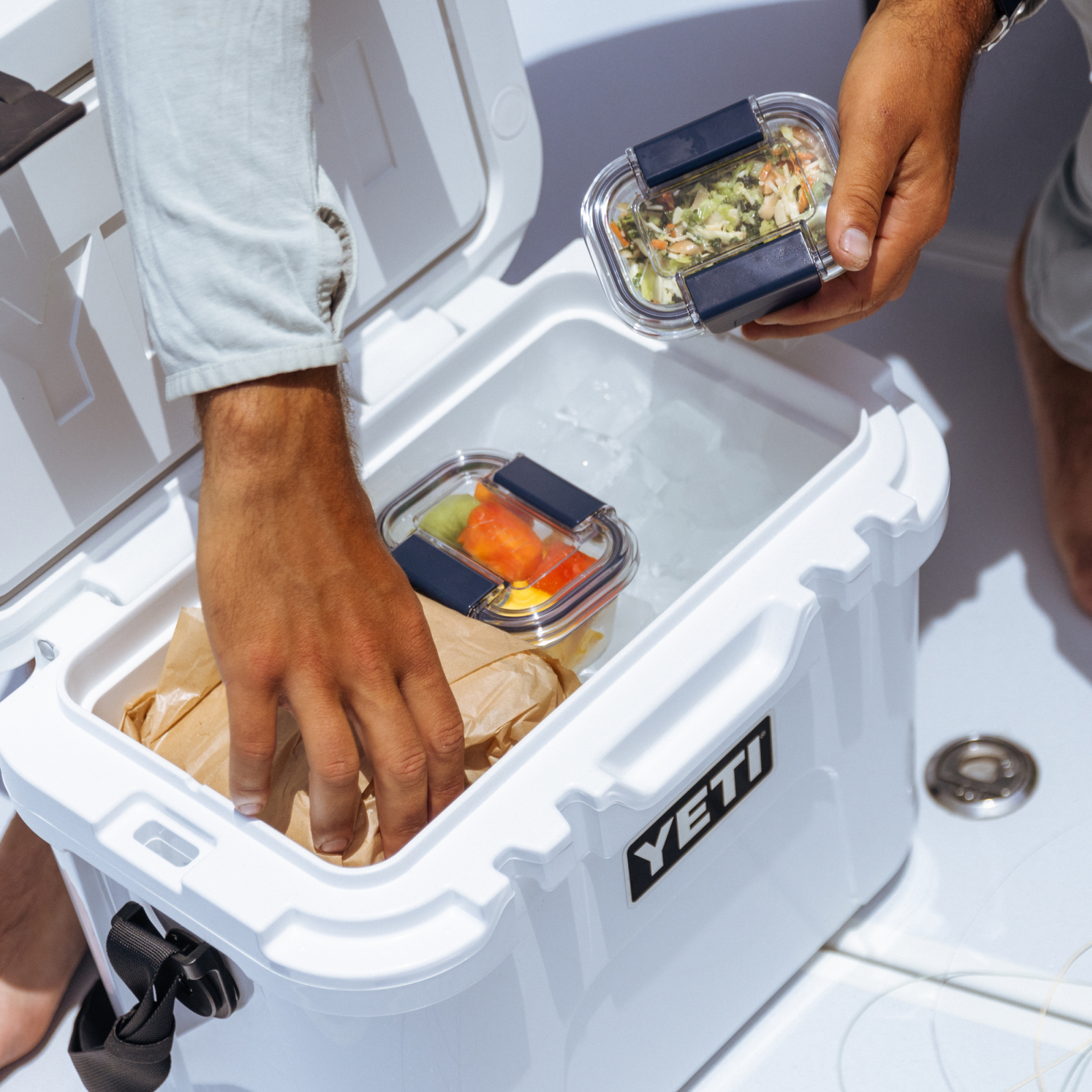 Zwei Personen packen eine weiße YETI-Kühlbox mit Eis, einen BPA-freien YETI® Food Storage Small Behälter und eine braune Papiertüte ein. Einer füllt Lebensmittel in die Kühlbox, während der andere einen auslaufsicheren Behälter mit Salat hält.