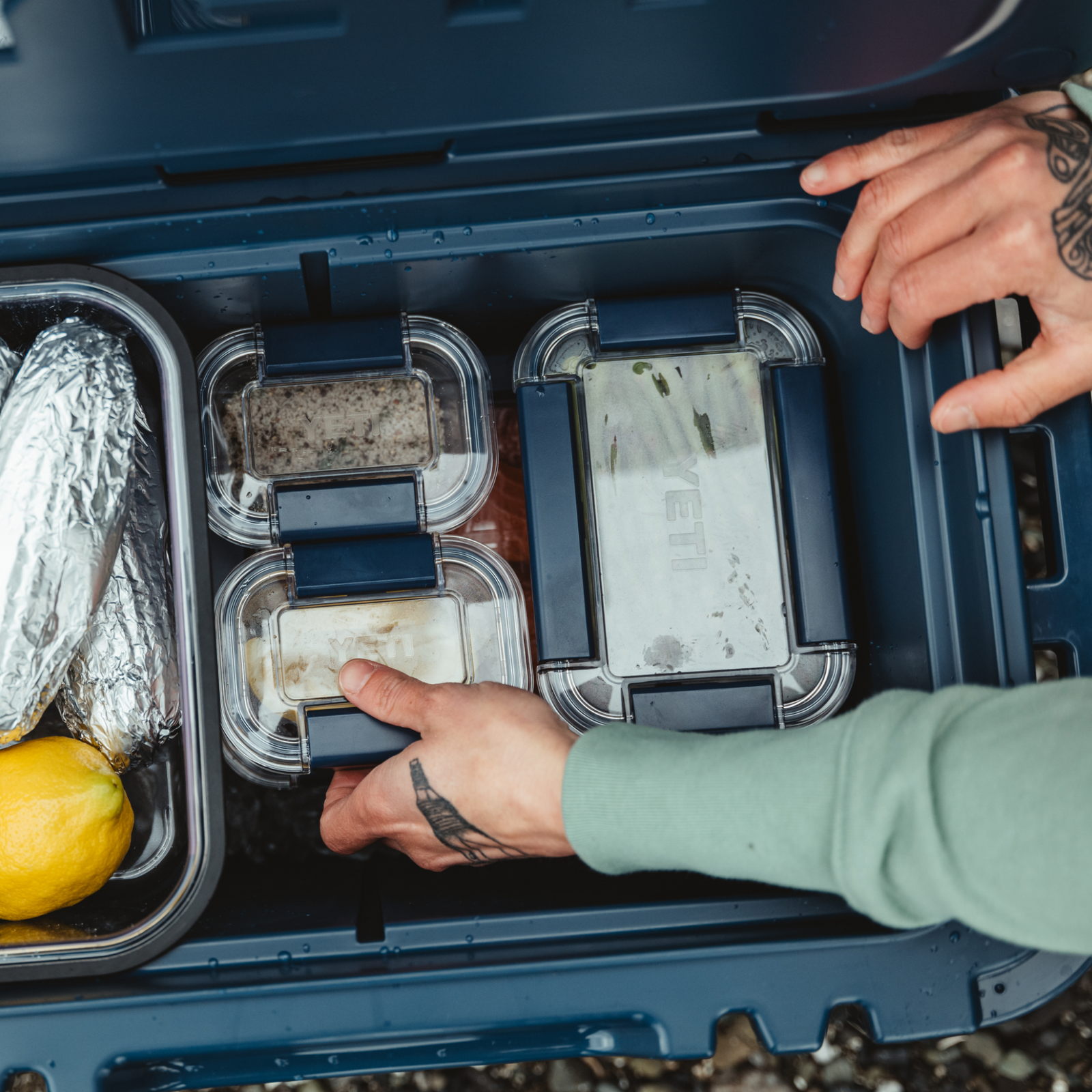 Eine Person mit Tattoos ordnet BPA-freie Lebensmittelbehälter, in Folie verpackte Snacks und Zitronen in einer blauen YETI® Food Storage Small Kühlbox von YETI.