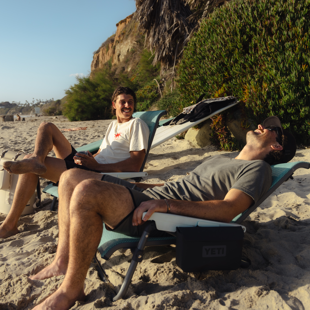 Zwei Männer entspannen und lachen an einem Sandstrand in der Nähe von Klippen und Grünflächen und genießen den sonnigen Tag, während sie auf ihrem YETI® Hondo Beach Chair - Seafoam sitzen.