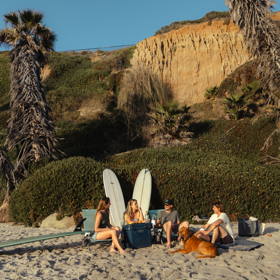 Vier Personen entspannen sich am Strand in YETI® Hondo Beach Chair - Seafoam Stühlen neben zwei Surfbrettern und einer Kühlbox und unterhalten sich mit einem braunen Hund. Palmen, grüne Sträucher und eine sonnenbeschienene Klippe bilden eine malerische Kulisse.