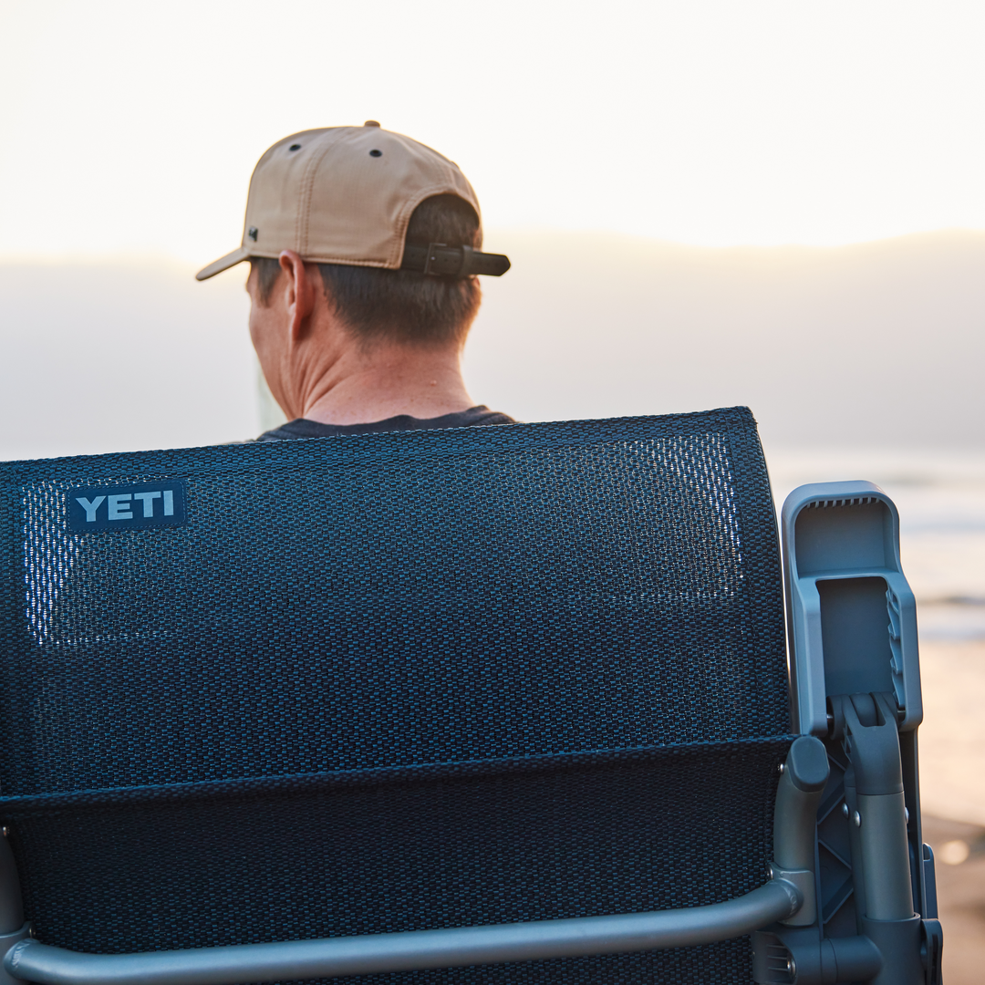Ein Mann mit hellbrauner Baseballmütze sitzt auf einem YETI® Hondo Beach Chair - Seafoam und blickt in den Sonnenuntergang über dem Meer. Von hinten betrachtet, schaffen der YETI-Stuhl und der ruhige Strand eine friedliche, entspannte Atmosphäre.