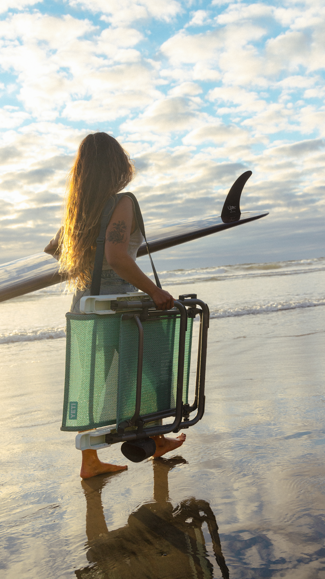 Eine Person mit langen Haaren läuft barfuß auf dem nassen Sand am Meer unter einem bewölkten Himmel, trägt ein Surfbrett und einen gefalteten YETI® Hondo Beach Chair - Seafoam von YETI.