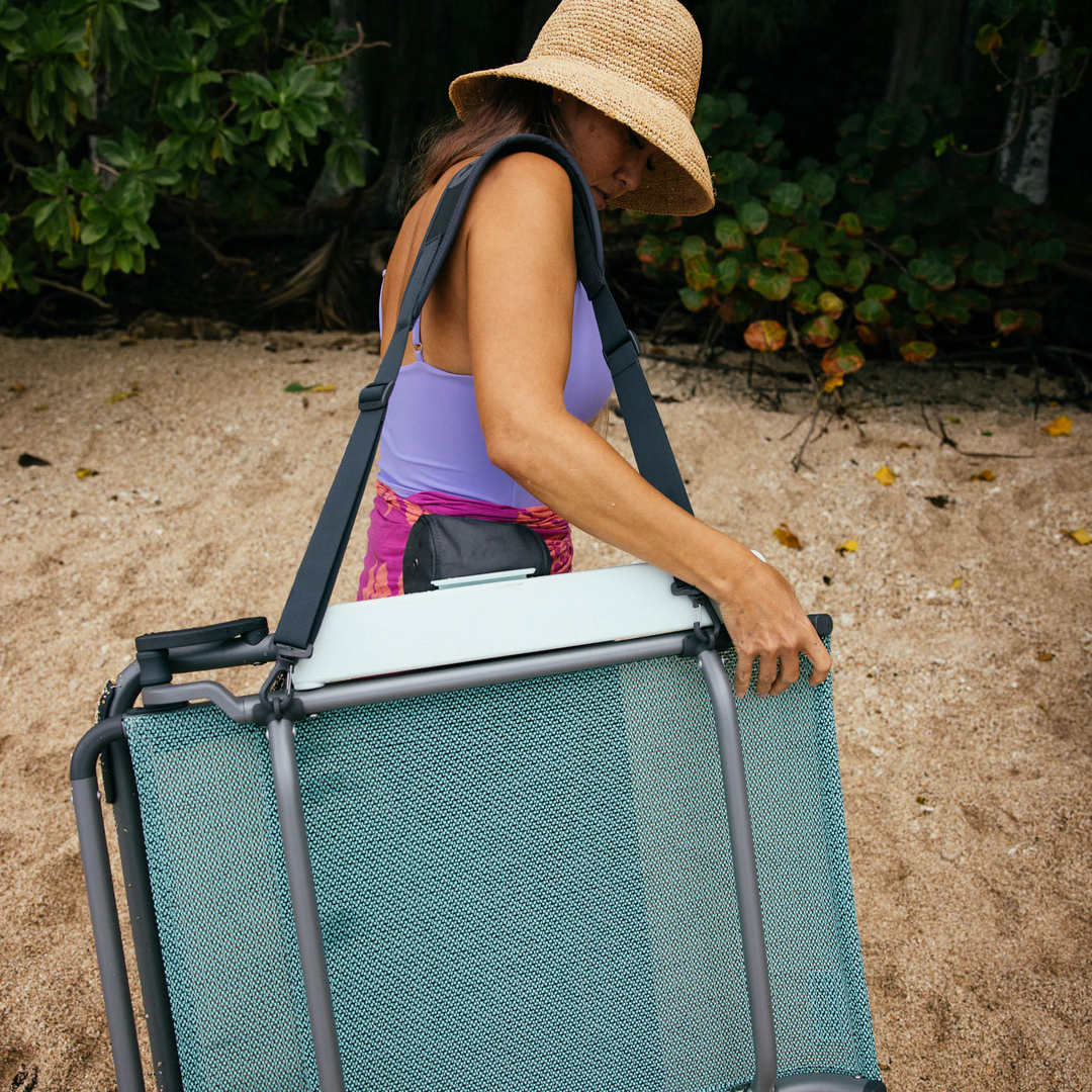 Eine Frau mit Strohhut und lila Badeanzug trägt einen gefalteten YETI® Hondo Beach Chair - Seafoam von YETI an einem Sandstrand, mit Grün im Hintergrund.
