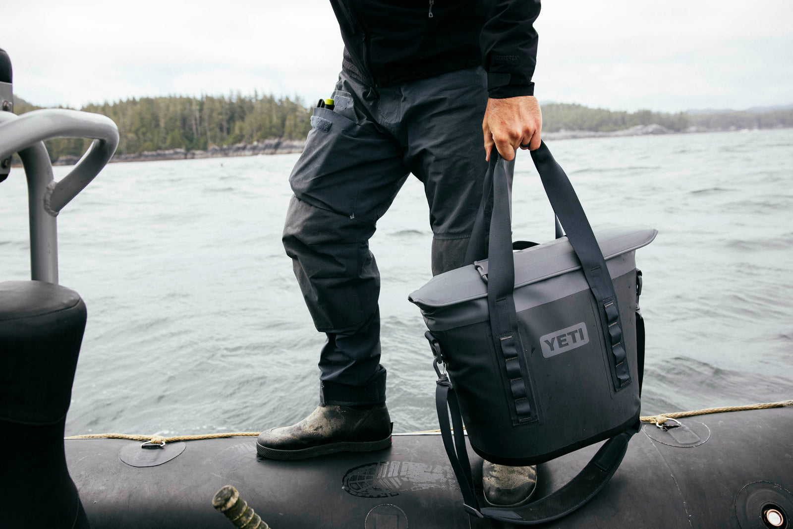 Eine Person in Outdoor-Ausrüstung steht auf einem Schlauchboot und hält eine YETI® Hopper M15 Soft Cooler - Charcoal an den Griffen. Im Hintergrund sind Wasser und eine bewaldete Uferlinie zu sehen.