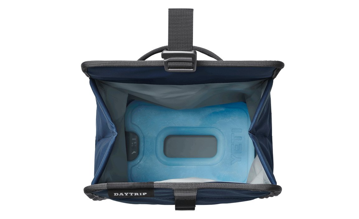 Draufsicht auf eine offene blaue Kühltasche mit der Aufschrift DAYTRIP, in der sich eine YETI® Kühlakku Ice Sheet Medium befindet, die Speisen und Getränke kühl hält. Die Tasche hat ein graues Innenfutter und einen Henkel an der Oberseite. Marke: YETI.