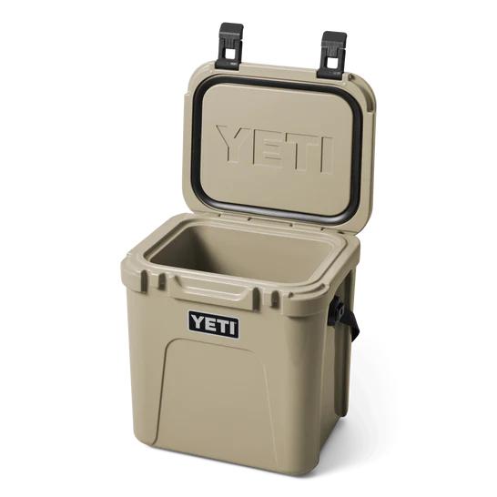 Eine hellbraune YETI® Kühlbox Roadie 24 mit geöffnetem Deckel zeigt einen leeren Innenraum, das eingeprägte YETI-Logo auf dem Deckel und ein Etikett auf der Vorderseite. Diese tragbare Kühlbox verfügt über robuste Seitengriffe und zwei schwarze Verschlüsse.