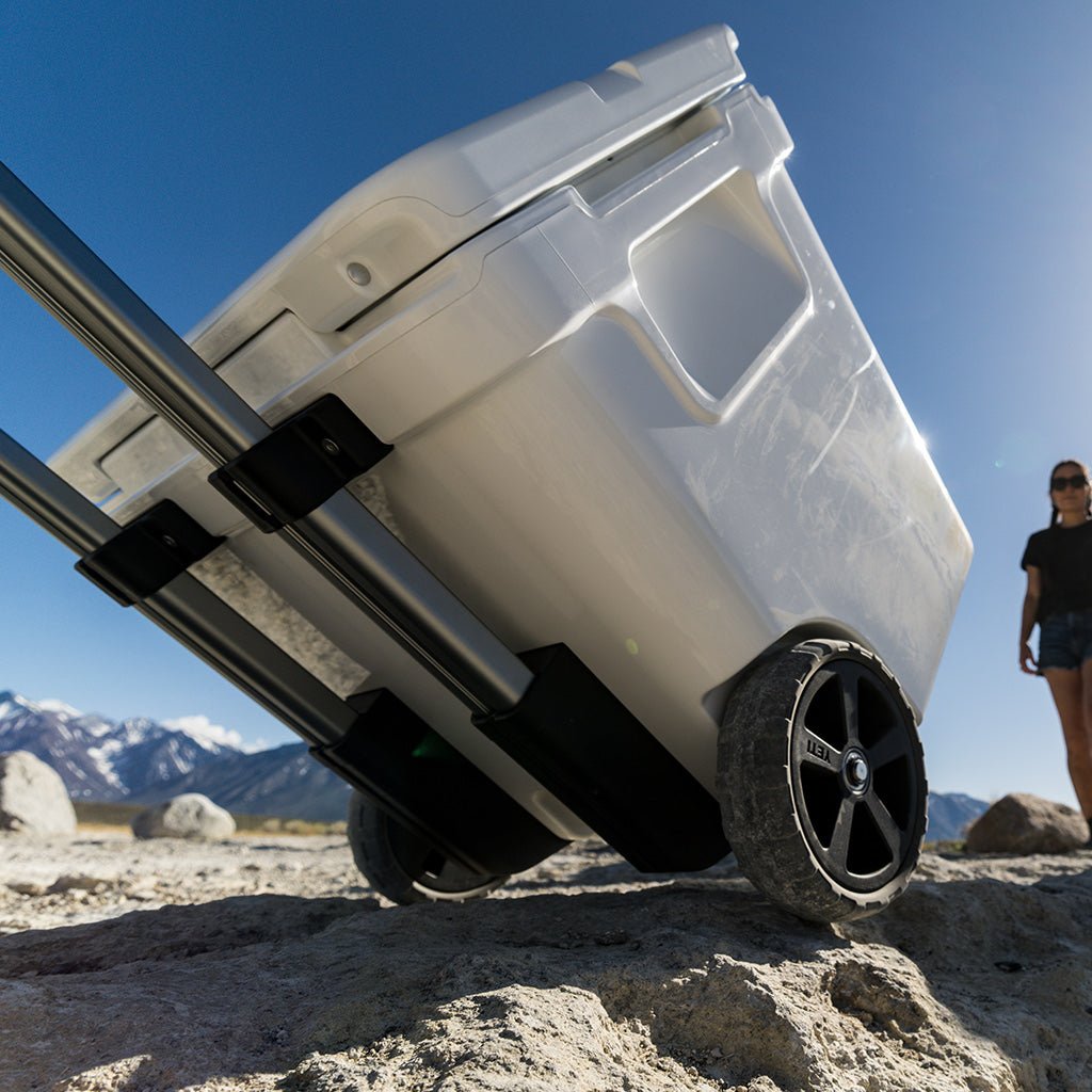 Eine Person zieht eine navyfarbene YETI® Kühlbox Roadie 48 mit Rädern und Teleskopgriff über felsigen Boden im Freien, mit Bergen und einem klaren blauen Himmel im Hintergrund.