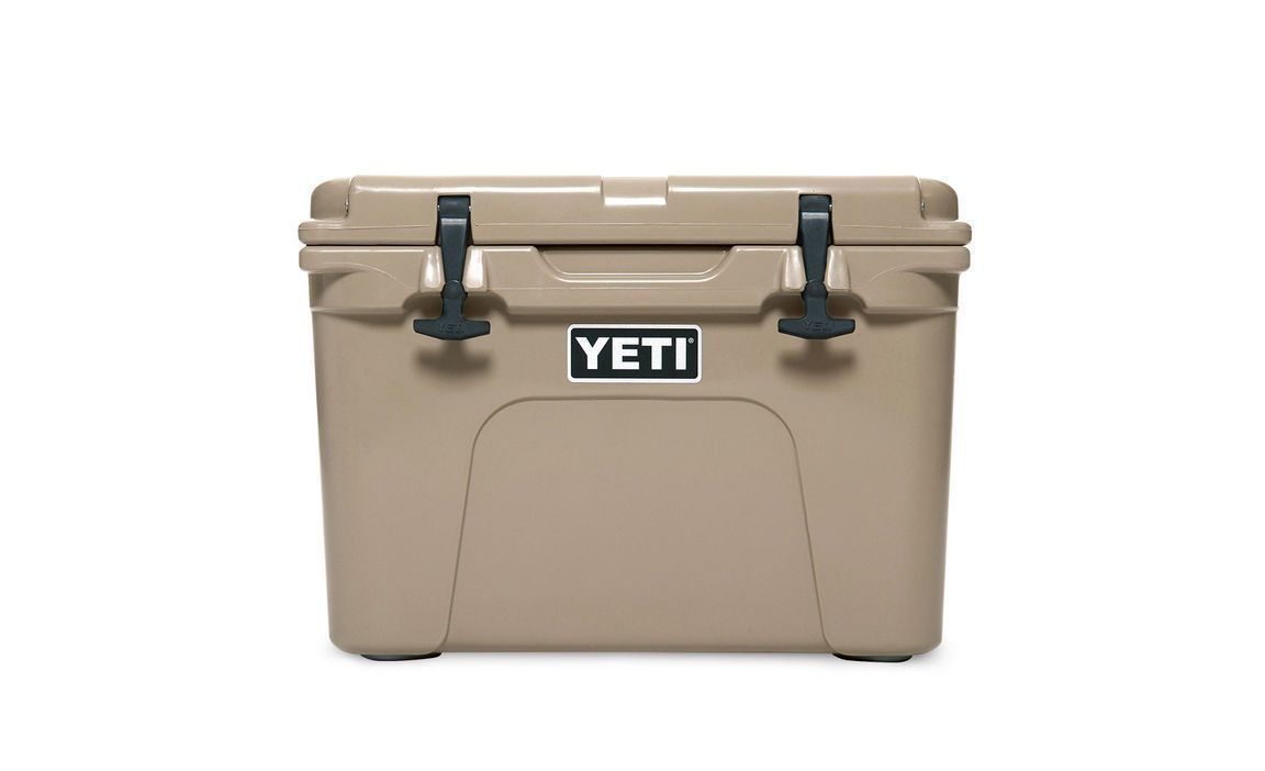Die YETI® Kühlbox Tundra 35 - Tan verfügt über zwei schwarze Frontverschlüsse und ein zentriertes YETI-Logo, das auf einem schlichten weißen Hintergrund abgebildet ist - ideal als robuste Kühlbox für jedes Camping-Abenteuer.