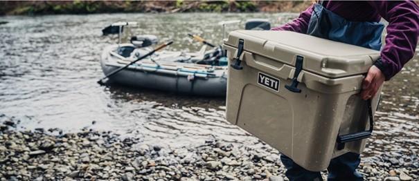 Eine Person steht an einem felsigen Flussufer und hält die YETI® Kühlbox Tundra 35 - White. Hinter ihr wartet ein Schlauchboot mit Rudern am Ufer, bereit für weitere Campingabenteuer mit dieser zuverlässigen YETI Kühlbox.