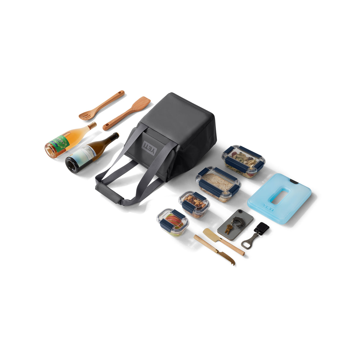 Ein Picknick-Set zeigt die YETI® Kühltasche Daytrip Lunch Bag 14L - Charcoal mit Coldcell Flex Isolierung, zwei Flaschen, Holzutensilien, vier Lebensmittelbehälter, blaues Schneidebrett, Flaschenöffner, Käsemesser, Reibe und Schäler auf weißem Hintergrund.